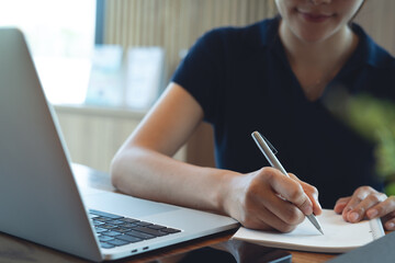 Business woman hand writing on notebook and research with laptop computer at home office. Freelancer online research, remote work and meeting agenda, business planning. Student studying online
