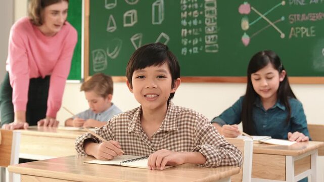 Asian smart boy smiling to camera while student writing answer in answer sheet. Multicultural student doing classwork or test while caucasian teacher checking student homework at classroom. Pedagogy.