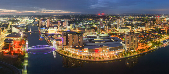 Fototapeta premium Night panorama of Salford Quays with illuminated bridges, glowing city lights, and vivid reflections on calm water.