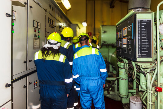 Seafarers in blue coveralls and yellow safety helmets are seen in the Emergency Generator Room of a merchant ship. They are engaged in operational training. 