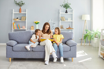 Mother and happy children are sitting on the couch at home, reading a book together. They are enjoying leisure time, sharing a family moment of fun and relaxation during the weekend.