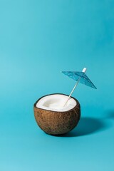 Minimal tropical composition with half a coconut filled with creamy drink and a small cocktail umbrella on a bright turquoise background, symbolizing summer relaxation, exotic vacation, and refreshmen