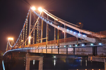 Beautiful night view of an illuminated suspension bridge over the river in with glowing lights, reflections on water, and impressive metal engineering design under dark sky