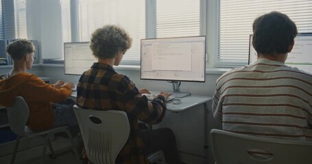 Group of Young University Students Working on Programming Tasks. Boys Coding Together in Bright Computer Lab. Focused Learners Type Code on Monitors, Studying Software Development. Back View. - Powered by Adobe