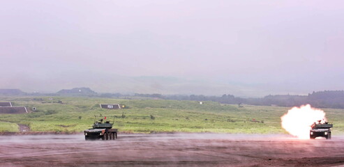 Amidst a foggy landscape, a powerful armored vehicle unleashes a bright muzzle flash, showcasing intense military training maneuvers across the expansive outdoor range with another tactical vehicle
