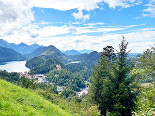 Panorama from above of Neuschwanstein Castle, hidden on the top of a hill surrounded by the mountain forests of the Bavarian Alps near Lake Alpsee against a blue, partly cloudy sky.