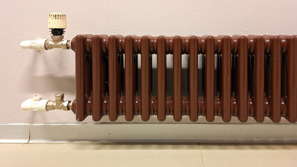 A long brown hot-water radiator against the wall. It has pipes and a temperature control. Close-up....