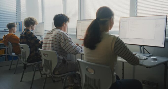 Young Students in Bright Classroom Learning Software Development. Focused Group Types Computer Code on Monitors, Exploring Programming, Teamwork, Digital Future Skills. Back View. Static Shot.