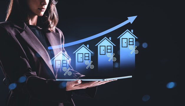 Woman using tablet with holographic rising house icons and percentage signs representing real estate growth concept on dark background..