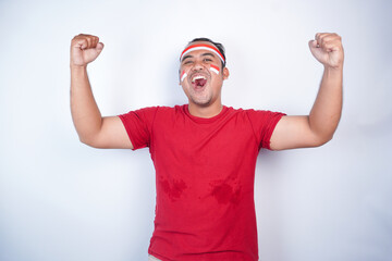 Naklejka premium A young Asian man with a happy successful expression, raised a fist wearing red shirt and flag headband isolated by white background. Indonesia's independence day concept.