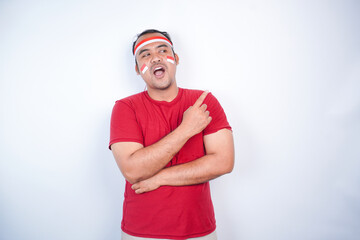 Fototapeta premium Shocked Asian man wearing red t-shirt, flag headband, pointing at the copy space on beside, isolated by white background. Indonesia's independence day concept.