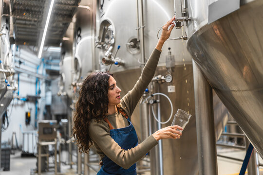 Woman brewer working in a modern craft beer brewery, checking beer quality by pouring sample from a fermentation tank - Powered by Adobe