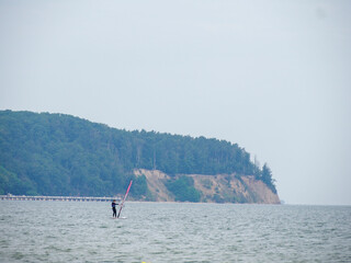 Wide shot of a stand-up paddleboarder on the sea, contrasted by a high forested cliff of the...
