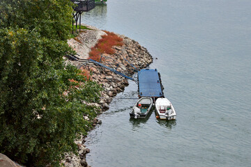 Two small boats moored at a blue floating dock on the rocky Danube River bank near Novi Sad, Serbia. Peaceful riverside view with greenery, autumn tones, and calm water under cloudy sky