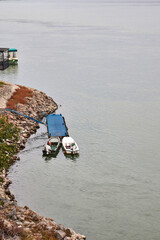 Two small boats moored at a blue floating dock on the rocky Danube River bank near Novi Sad, Serbia. Peaceful riverside view with greenery, autumn tones, and calm water under cloudy sky