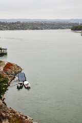 Two small boats moored at a blue floating dock on the rocky Danube River bank near Novi Sad, Serbia. Peaceful riverside view with greenery, autumn tones, and calm water under cloudy sky