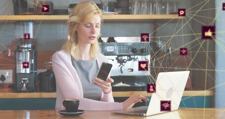Typing woman wearing pink cardigan on silver laptop in coffee shop, with smartphone, network icons