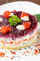 Elegant plated Russian layered salad with beetroot, herring, mayonnaise, red caviar, and parsley garnish on white plate over wooden table. Gourmet presentation, top view.