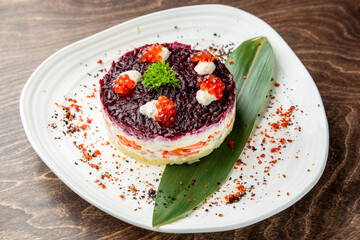 Elegant plated Russian layered salad with beetroot, herring, mayonnaise, red caviar, and parsley garnish on white plate over wooden table. Gourmet presentation, top view.