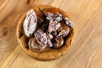 Close-up of a caramel cookie topped with glazed pecan and sugared nut pieces on a wooden surface.