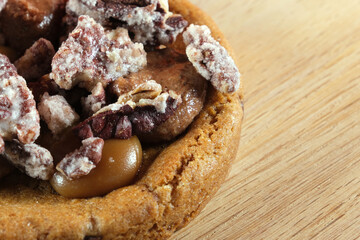 Close-up of a caramel cookie topped with glazed pecan and sugared nut pieces on a wooden surface.