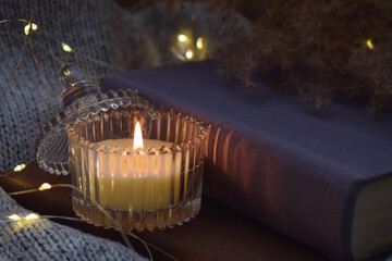 Candle on closed book and cozy knitted fabric with warm fairy lights creating peaceful evening home atmosphere.