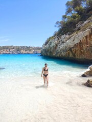Young slim woman in black bikini comes out of sea, walk white sand beach, turquoise water and blue sky in background. Active lifestyle, carefree vacation. Solo Model photoshoot. Natural beauty. Blog