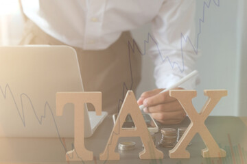 Tax, investment, business planning, and economic stock chart analysis concept with person working on laptop and calculator beside stacked coins and large TAX letters on desk