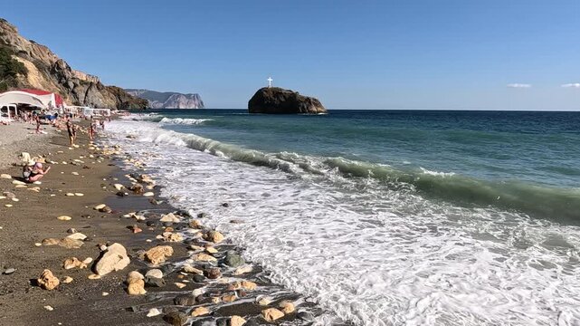 Beach waves sea, people relaxing on a pebble shore with crashing waves and a distant island under a clear summer sky