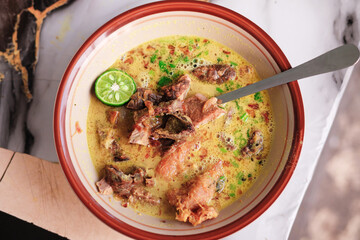 A top-down view of Soto Kuning Bogor, a traditional Indonesian yellow beef soup. The savory broth is garnished with a lime slice and fresh herbs, with a spoon resting in the bowl. Bogor popular Soup