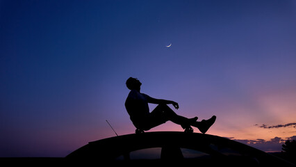 Man watching stars, Milky Way, planets, Moon and other celestial objects from a car rooftop from a dark countryside location.
