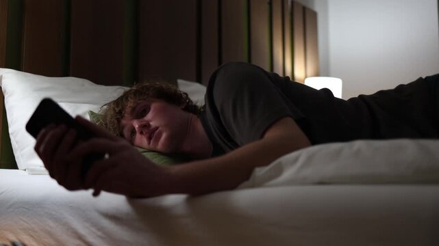 Young man lying in bed at night scrolling through smartphone people spending time online instead of sleeping symbolizing digital addiction modern lifestyle and restless insomnia illuminated by soft li