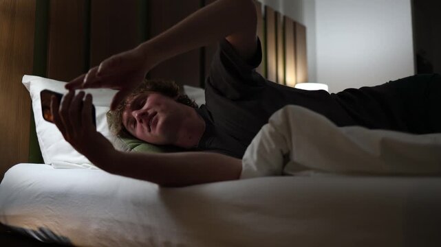 Young man lying in bed finally puts down his phone and falls asleep peacefully showing relaxation digital detox and relief in cozy dimly lit bedroom with calm evening atmosphere