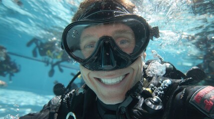 A diver with a black mask and equipment smiles widely underwater in a swimming pool. Bubbles surround him as other divers practice in the background enjoying their training.