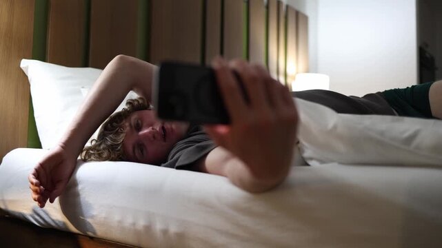 Teen boy lying in bed late at night taking selfie with smartphone showing social media obsession and youth addiction to online attention in dark cozy room with phone light glow