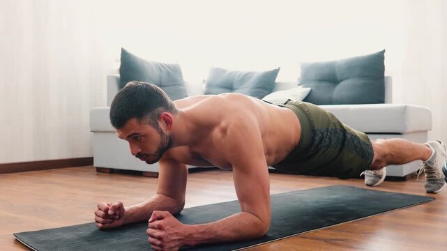 Young man exercising at home. Video of strong powerful guy stand in plank position on yoga mat. Confident concentrated man training in living room.