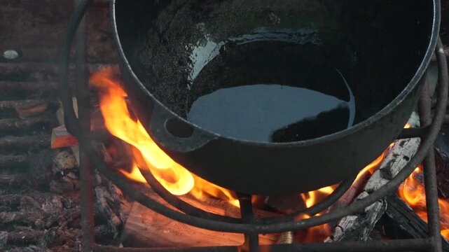 Fire cooking pot, old cast iron with oil heats over roaring campfire, chopped carrots added outdoor traditional cooking