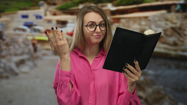 Woman in pink shirt reading book at beach, gesturing money sign, with blurred seaside background, conveying financial independence and relaxation outdoors.