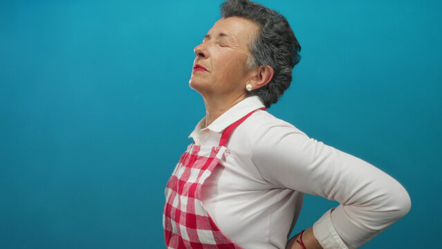 Senior woman with grey hair wearing an apron stands against a blue background, holding her back in discomfort, suggesting back pain and age-related issues.