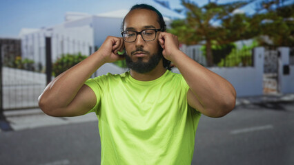 Man plugs ears with fingers on street at house entrance, wearing bright green shirt and glasses,...