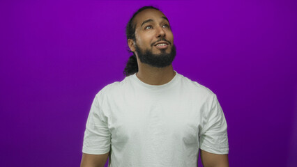 Man smiling with visible teeth and beard looking up in studio with purple backdrop wearing white t...