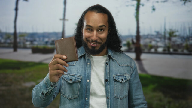 Man wearing denim jacket and white t shirt holding a brown wallet and smiling while looking down on a street by a marina; casual joy.