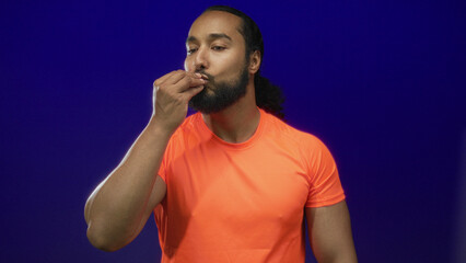 Man gesturing with raised hands and open palms toward camera in blue studio wearing bright orange...