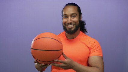 Man holding basketball with hands in purple studio wearing orange shirt, muscular biceps visible and smiling; confidence.