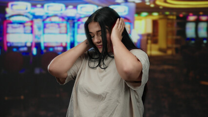Woman with closed eyes and hands covering ears stands in a bustling indoor casino next to vibrant slot machines, showcasing a serene yet immersive atmosphere.