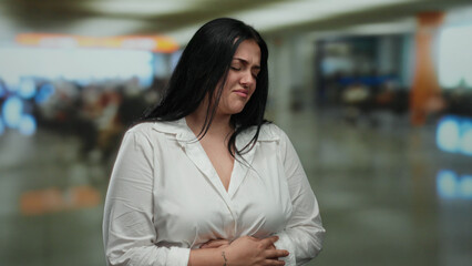 Fototapeta premium Woman in white shirt with stomach pain at bustling airport terminal with blurred background showcasing discomfort in public setting.