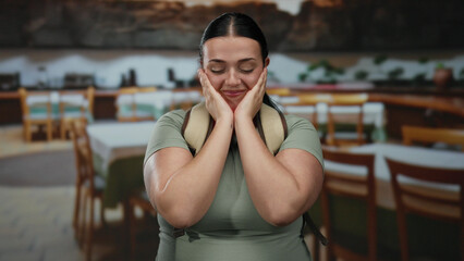 Woman smiling indoors in a restaurant setting, wearing casual attire and a backpack, capturing a light-hearted moment of a plus-size, young female enjoying coffee ambiance.