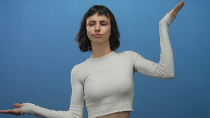 Young hispanic woman wearing white crop top holds palms up and looks sideways in blue studio; doubt...