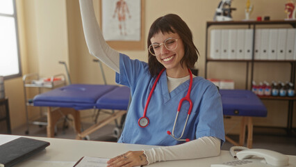 Obraz premium Woman nurse wearing blue scrubs and stethoscope raises arm in building; confidence empowerment success.