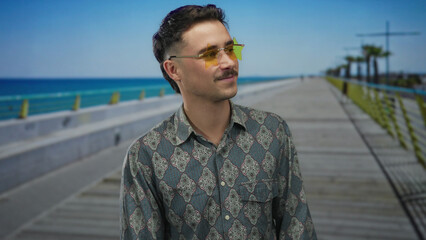 Young hispanic man with sunglasses standing on a seaside promenade with a patterned shirt under a clear blue sky.